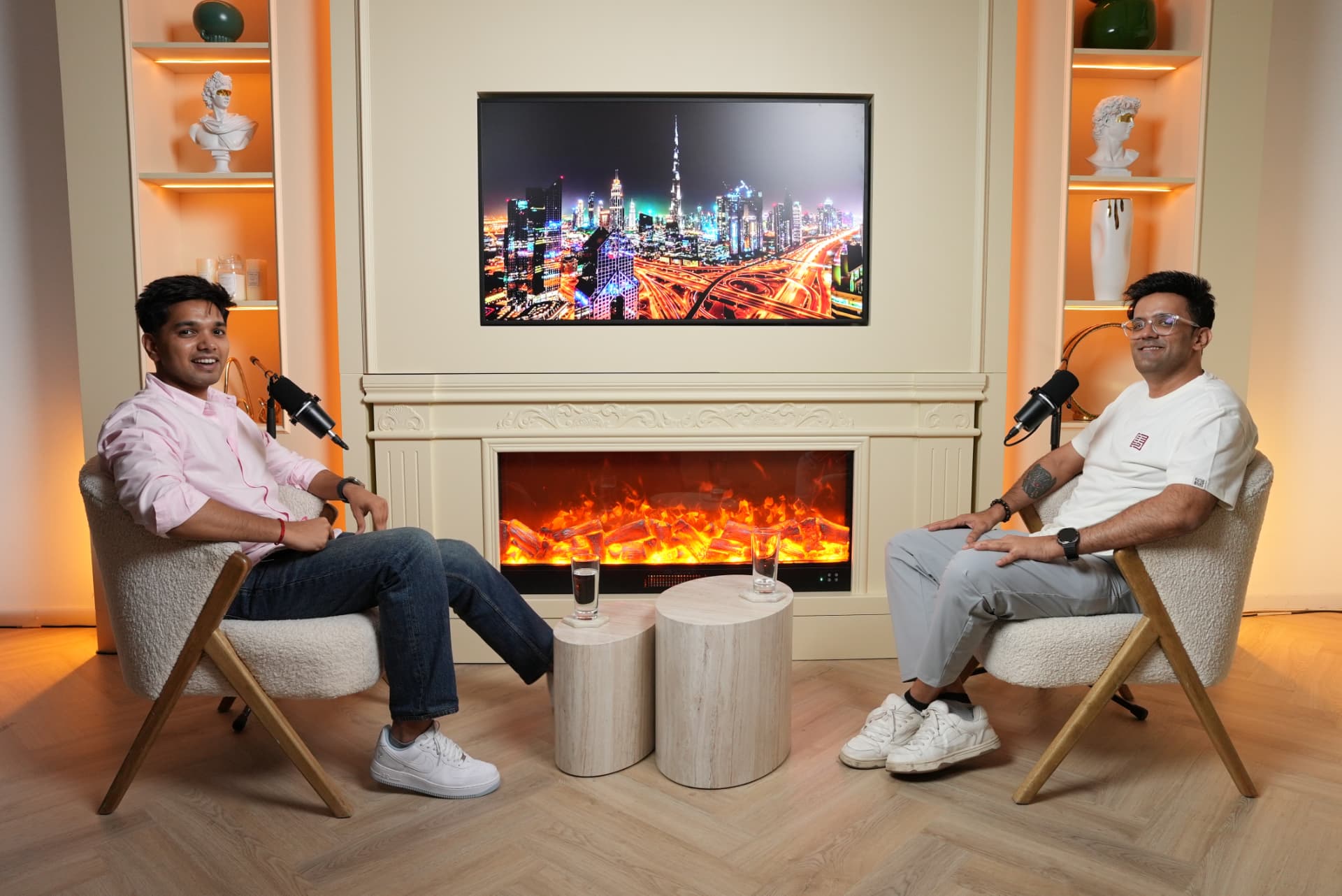 Two people seated in cream armchairs facing each other with broadcast microphones in a studio lit by a fireplace